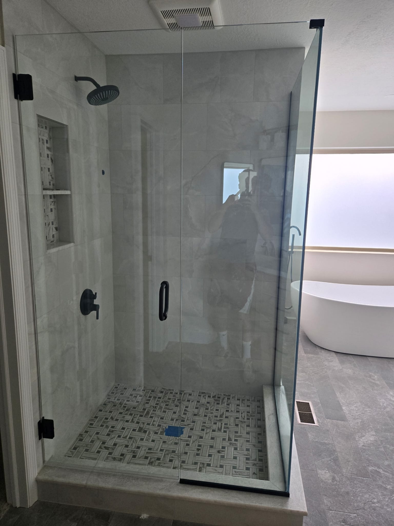 Walk in shower with marble look porcelain walls, basketweave mosaic floor, and tiled niche, finished with custom glass through our glass partner